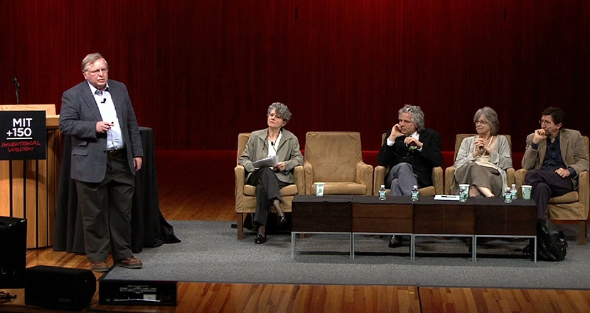 A man stand beside podium on stage, looking at the row of four seated panelists on stage, red curtain in background.