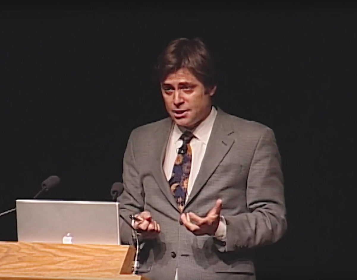 Max Tegmark speaks onstage, a podium and black curtain in the background