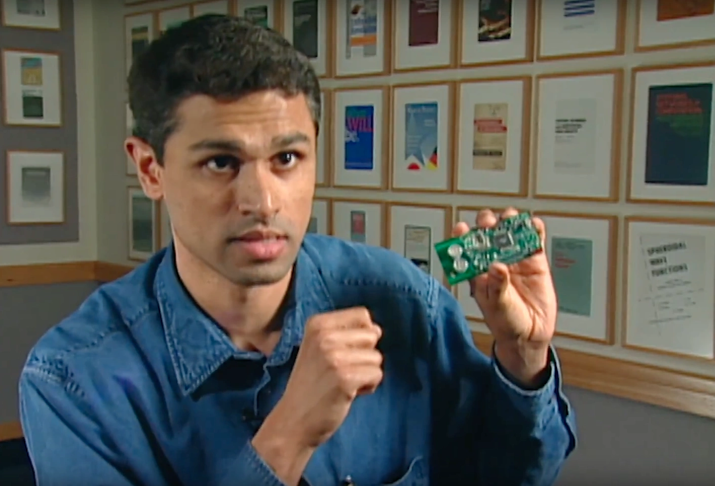 Man wearing blue shirt holds up small technological device. Rows of framed photos line the walls behind him.