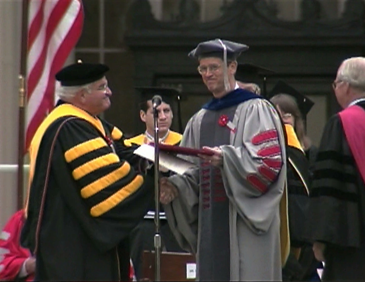 Charles M. Vest and Paul E. Gray, both wearing commencement attire, shake hands on a stage outside.