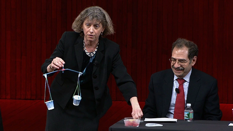 Hammel on stage holding up homemade model of balancing scales, Marc Kastner at panelist table in background.