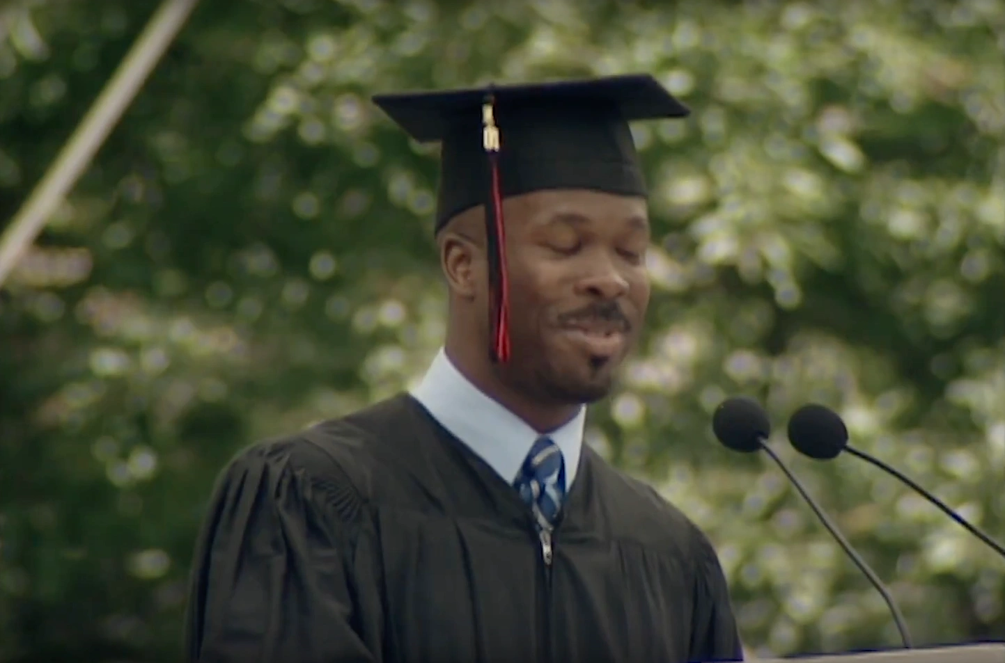 Elias A Zerhouni wearing a black cap and gown, speaking at podium on outdoor stage, trees in background.