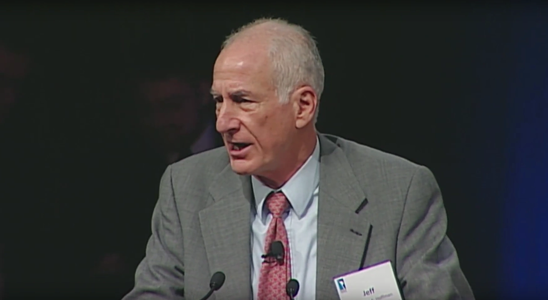 Joseph G. Gavin Jr. speaking at podium on stage, blue curtain in background