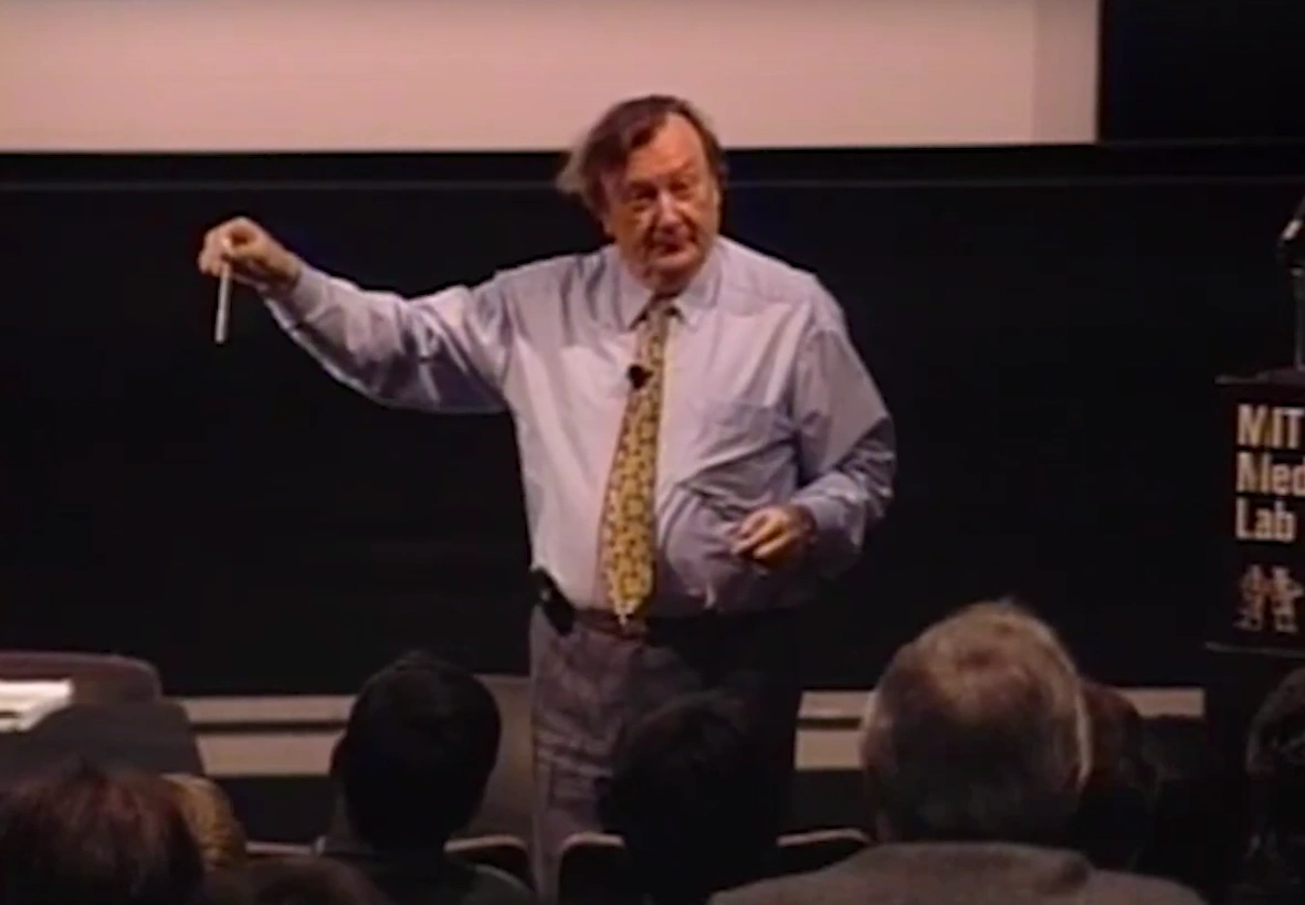 Carlo Rubbia speaking on stage, one arm extended to his side, he holds pen-shped object.  