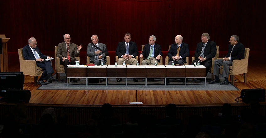 A panel of MIT alumni are seated in chairs on stage for a discussion, red curtain in background.