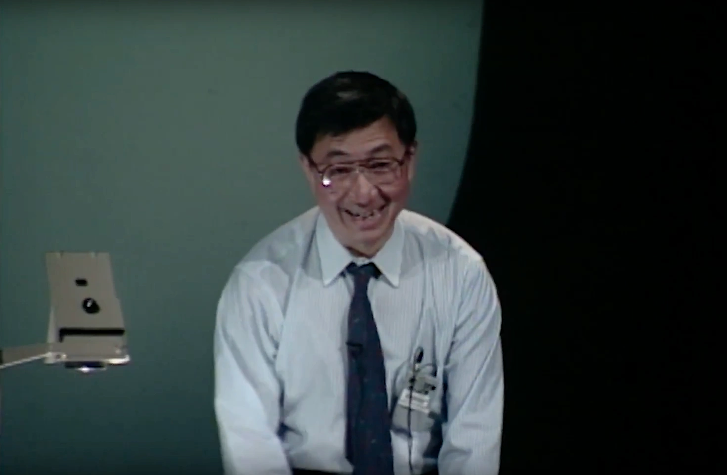 Samuel C. C. Ting stands on stage beside an optical microscope, smiling. Green wall in background.