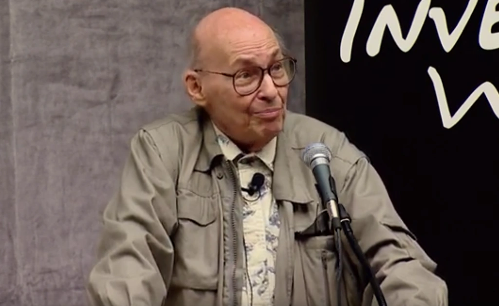 Marvin Minsky speaks into a microphone onstage, a dark backdrop in background.