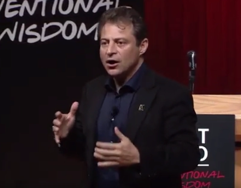 Peter Diamandis speaks on stage walking in front the podium, a dark backdrop in background.
