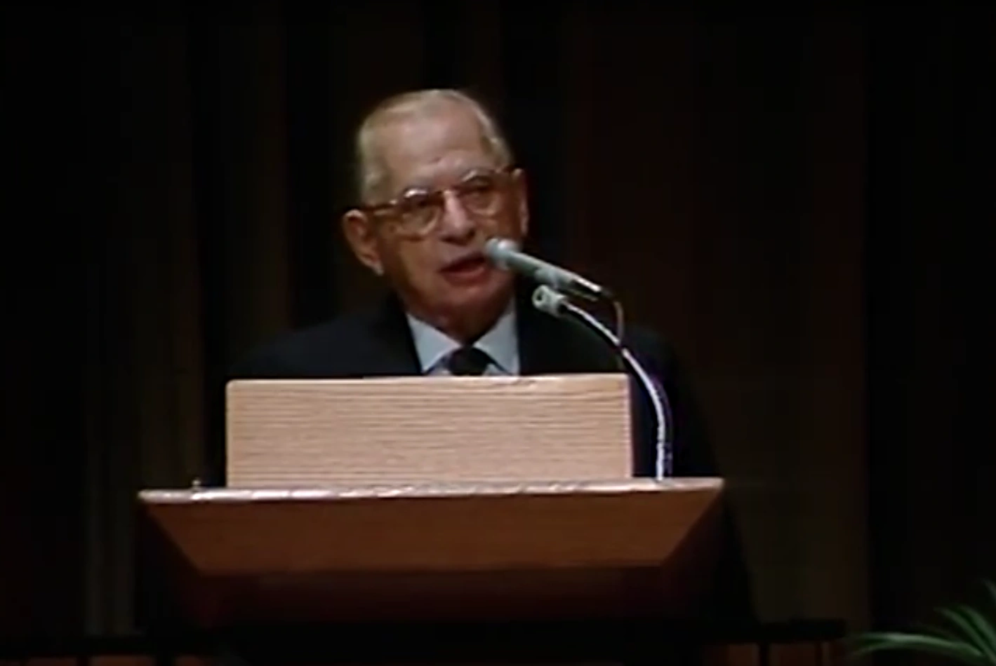 Ralph Landau speaks into podium on stage, black curtain in the background