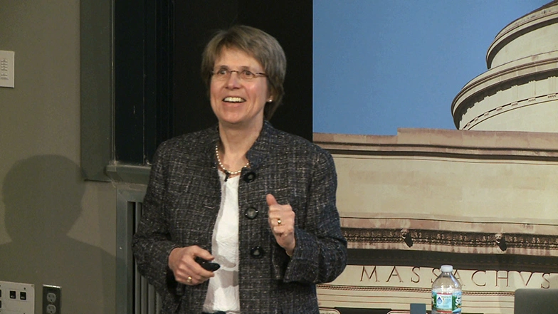 Sallie Chisholm gives speech in classroom, an MIT Great Dome backdrop is partially covering a chalkboard in background.