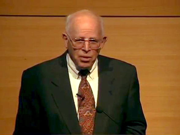 John H. Gibbons speaking on stage in front of a woodpaneled background