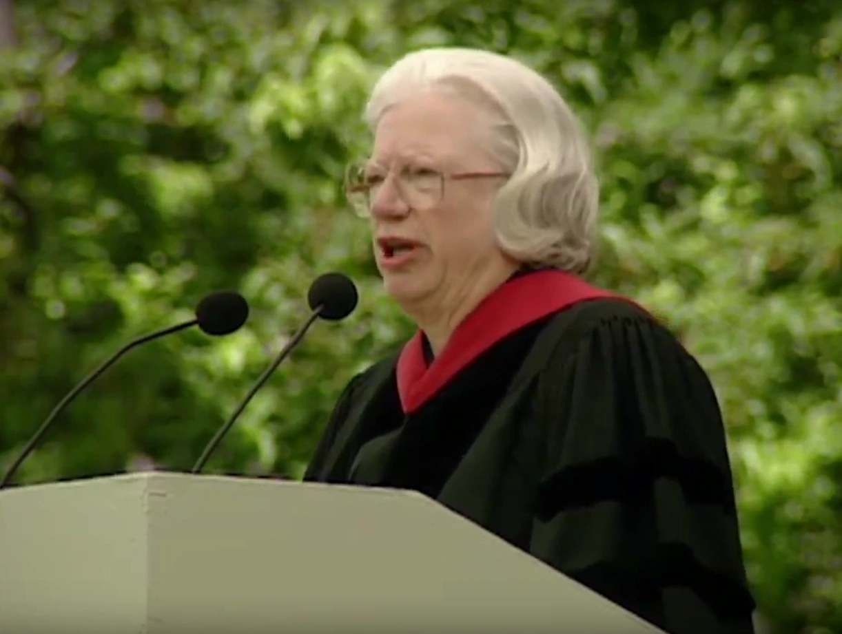 Hanna Gray, wearing commencement attire, speaking at podium on outdoor stage, trees in background